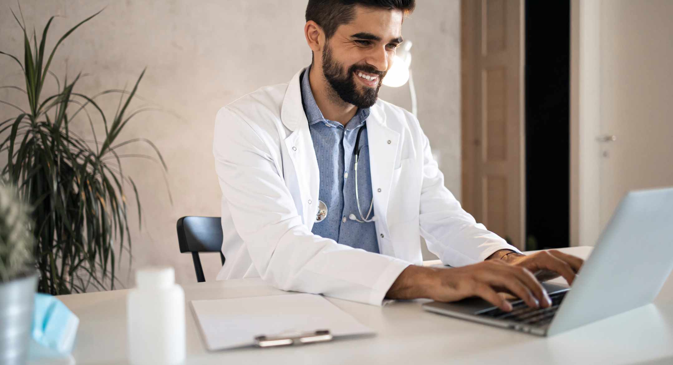 A doctor smiling at his desk while he types on his laptop