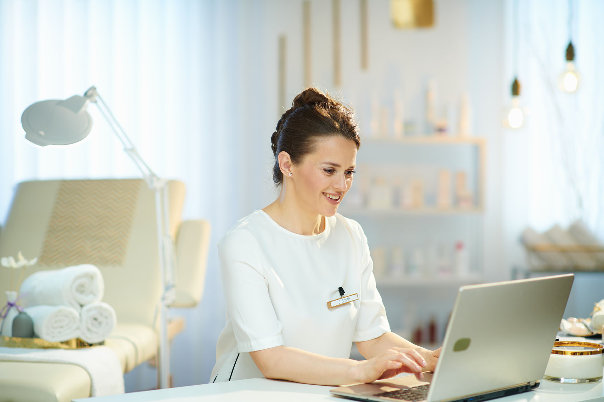 A female dermatologist sitting in a medical patient room, she's wearing a white outfit and sitting down typing on a laptop