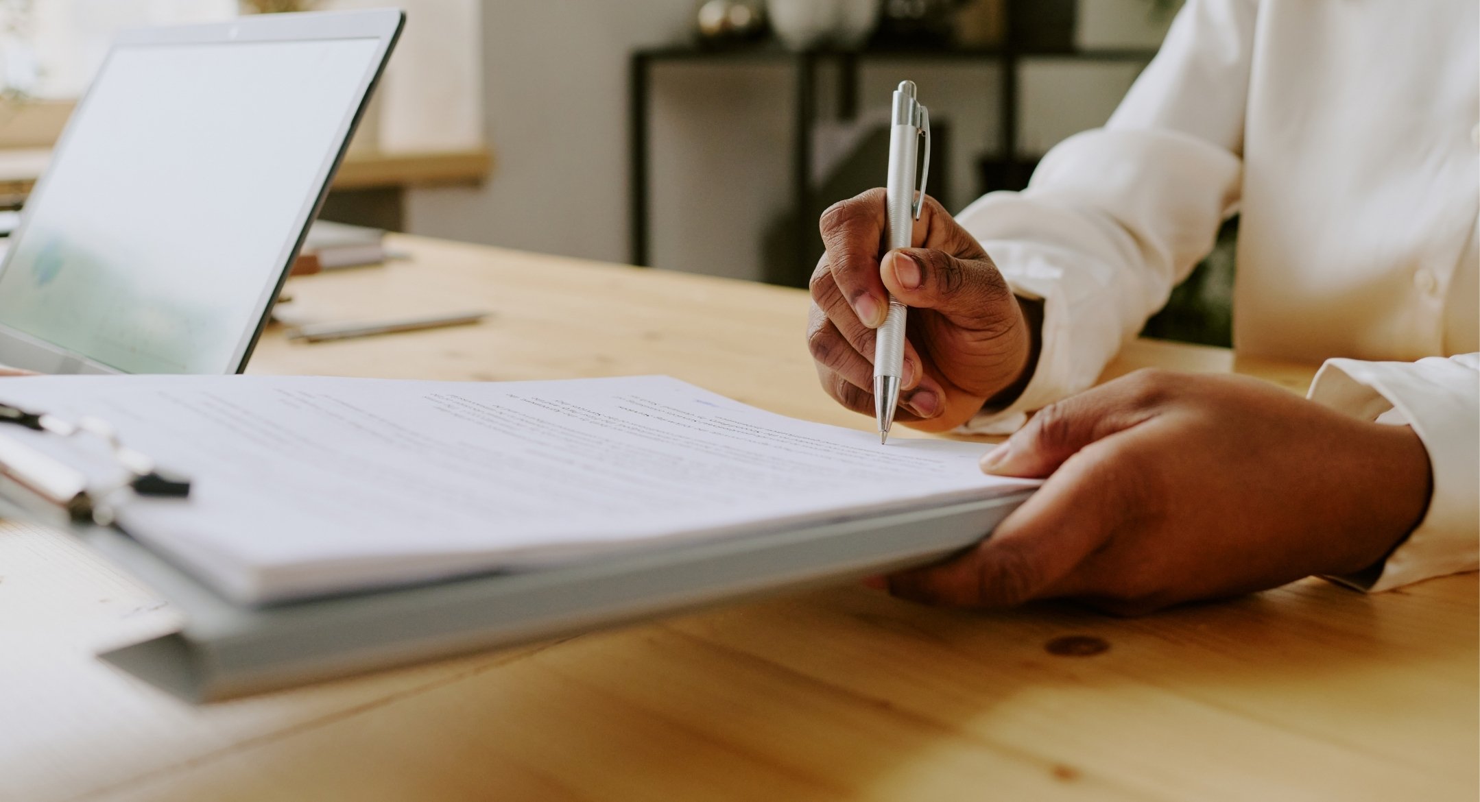 womans hand holding and signing on pile of papers with laptop on the side, indoor office room