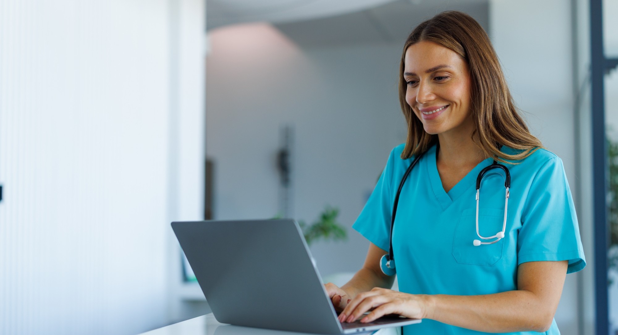 A clinician in scrubs smiles while typing on a laptop